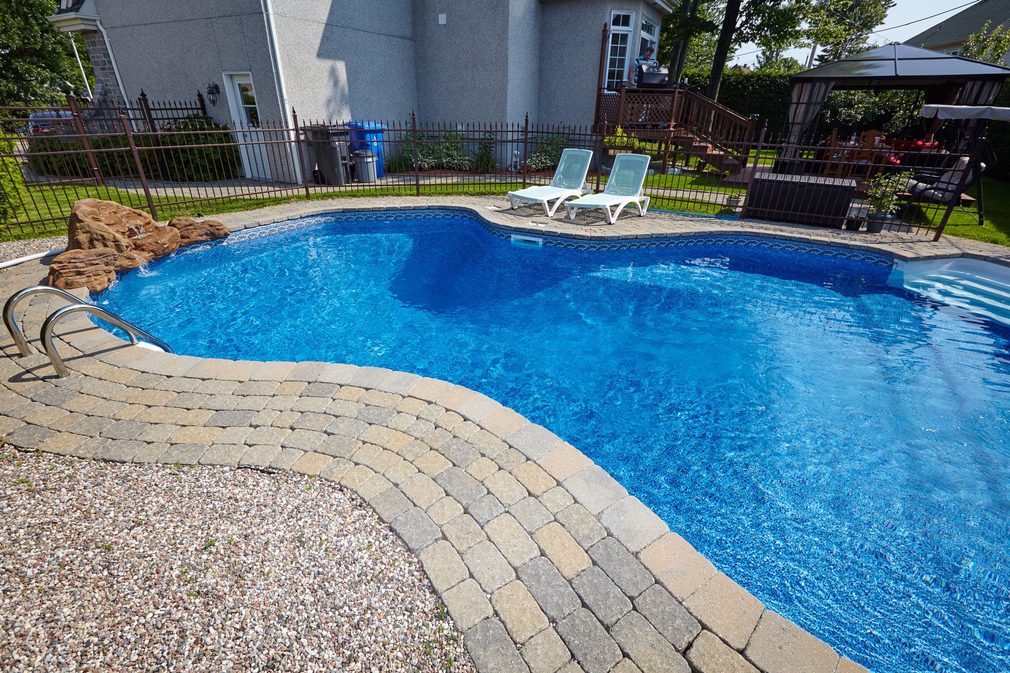 shutterstock_530860597 A backyard with a kidney-shaped swimming pool, two white lounge chairs, a shaded seating area, and surrounding greenery.